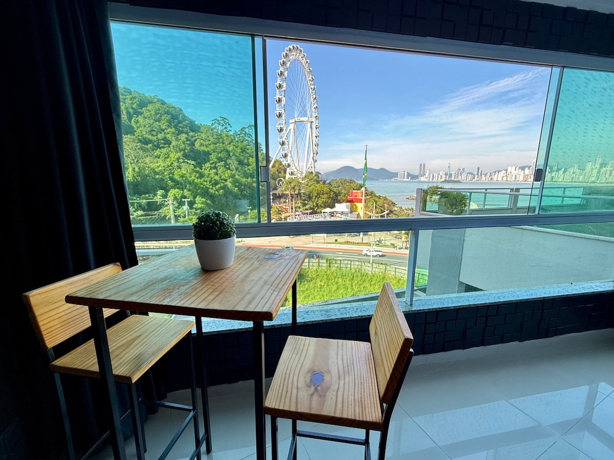 A dining area features a wooden table with two matching chairs. Large glass windows offer a panoramic view of the beach, the Ferris wheel, and the surrounding landscape. Natural light brightens the space, enhancing the connection to the outdoors.