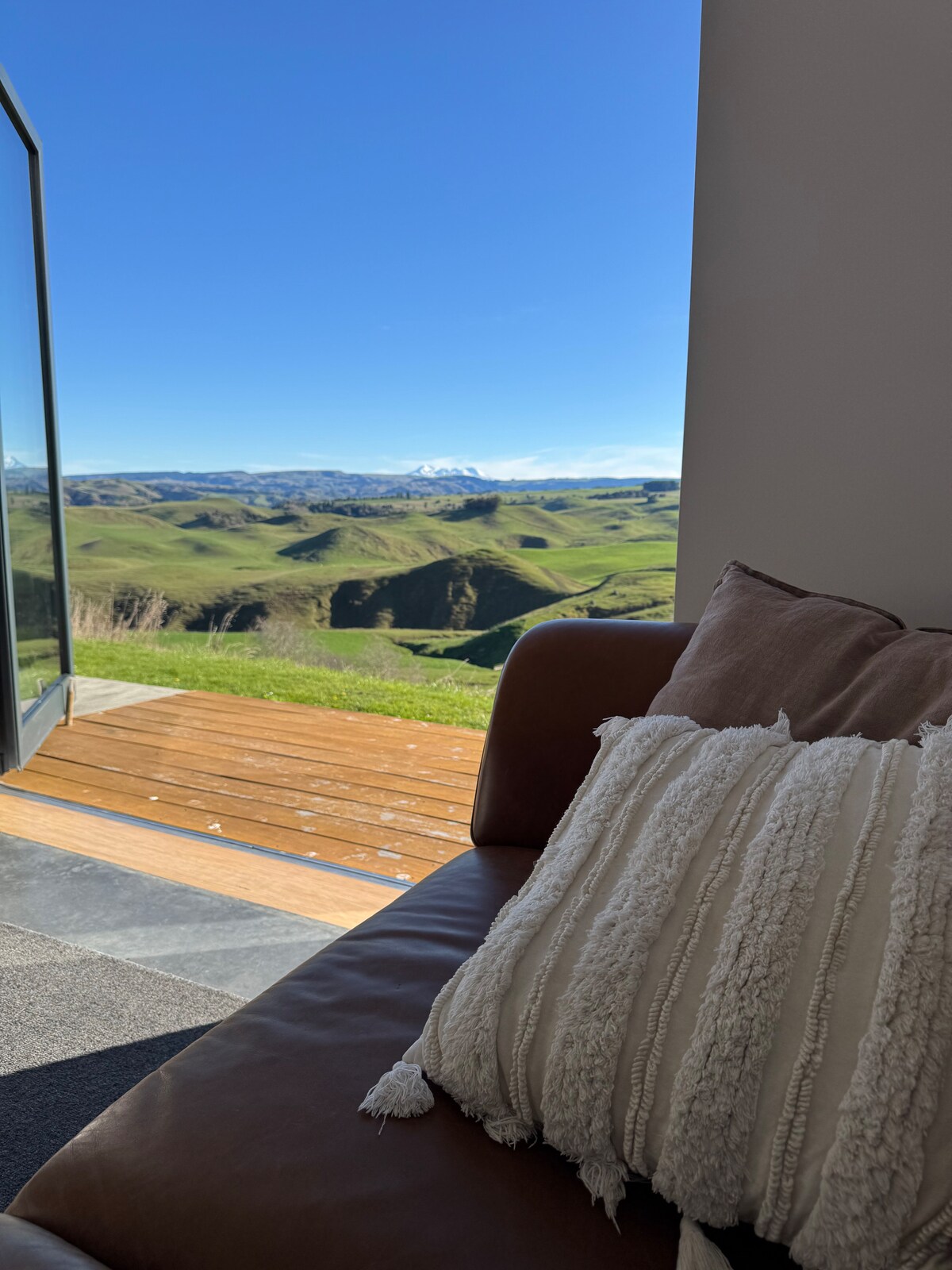 A cozy seating area features a brown leather couch adorned with a textured white pillow. Floor-to-ceiling glass doors open to reveal expansive views of rolling green hills under a clear blue sky, with distant snow-capped mountains visible on the horizon.