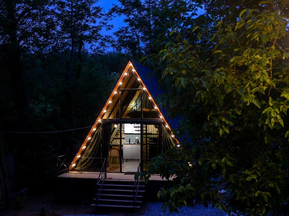An A-frame cottage is illuminated by string lights, highlighting its unique triangular shape. The structure features large windows that reflect the surrounding trees, and a spacious porch is visible, providing access to an inviting entrance.