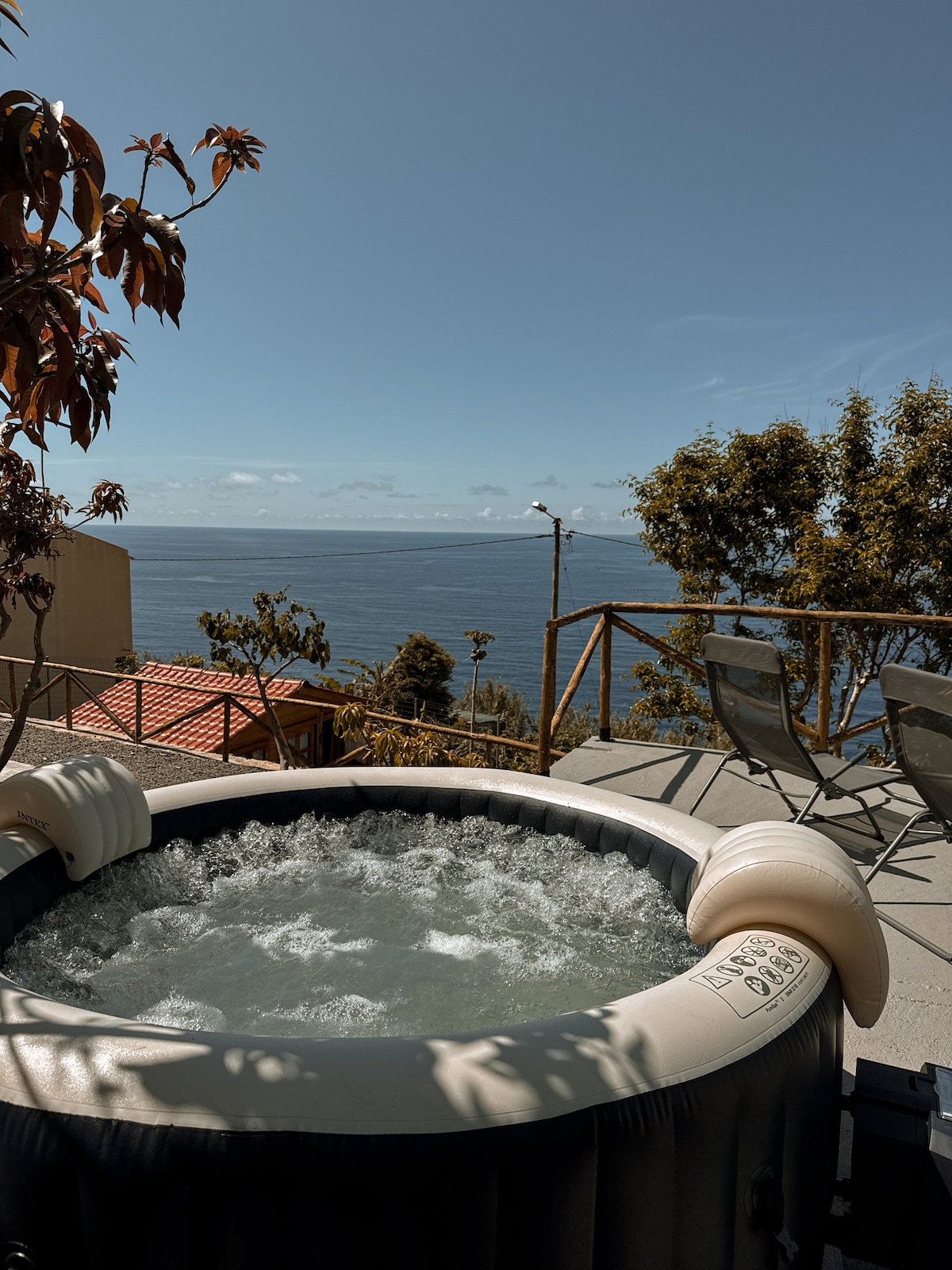 A spacious jacuzzi is situated on a sunlit terrace, offering uninterrupted views of the ocean. The bubbling water contrasts against the serene blue of the sea. Nearby, two chairs are positioned, inviting moments of relaxation amidst a natural setting.