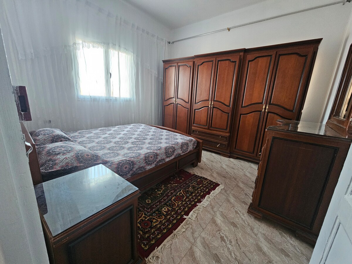 A cozy bedroom features a spacious bed with an intricate patterned bedspread. A wooden wardrobe and a matching dresser complement the warm wood tones. Natural light enters through a window with sheer curtains, illuminating the room softly. A decorative rug lies on the tiled floor.