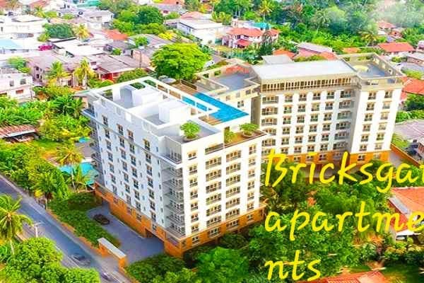 An aerial view of a modern apartment complex is presented, showcasing a rooftop pool area amidst lush greenery. The building features multiple stories with balconies and landscaped terraces, positioned within a vibrant neighborhood filled with trees and residential buildings.