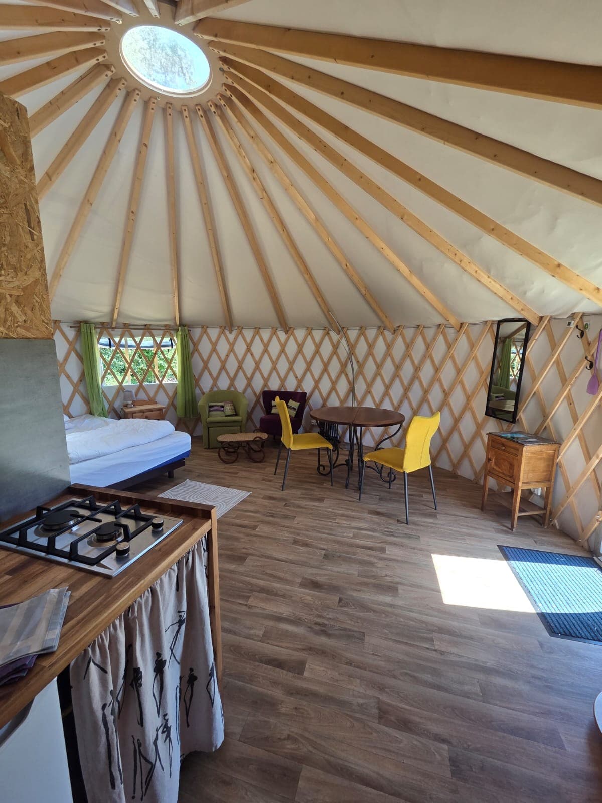 The interior of the yurt showcases a spacious layout with light wood accents and ample natural light from a circular skylight. A dining table with yellow chairs is positioned near a window, while a cozy sleeping area is visible in the background.