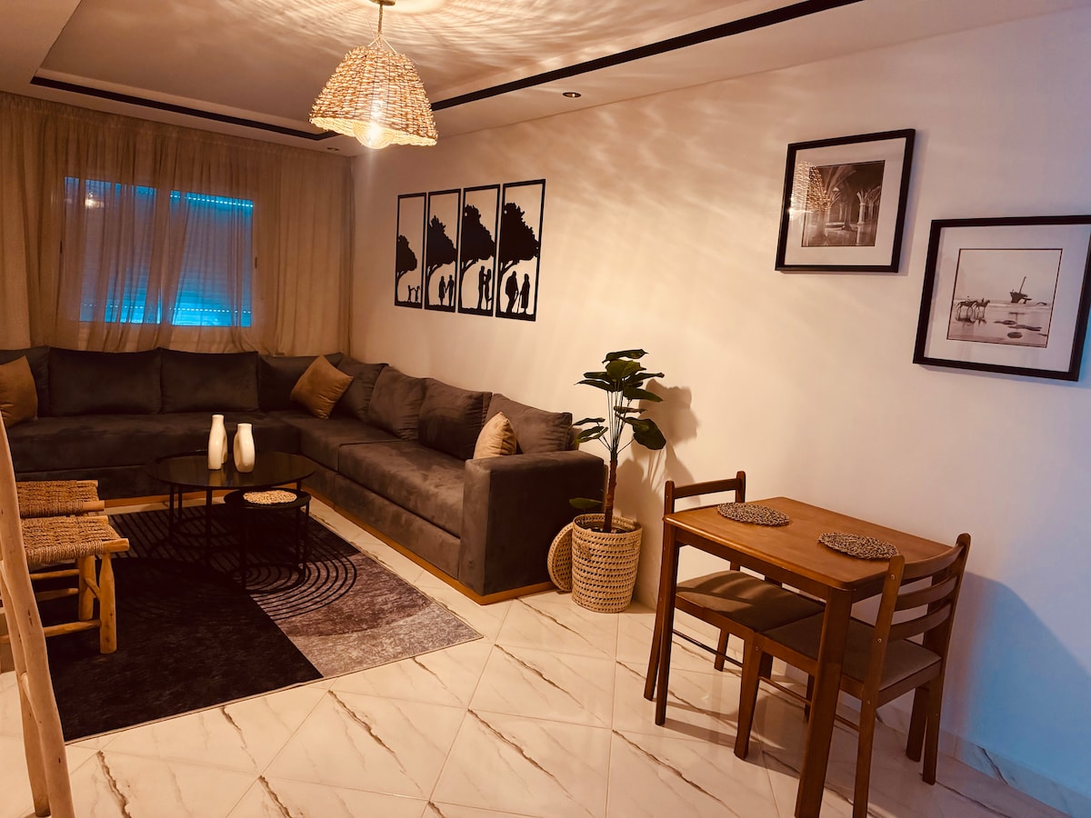 A cozy living area features a large gray sectional sofa adorned with decorative cushions. A circular coffee table is centered on a textured rug, complemented by a simple dining arrangement with two wooden chairs and a small table. Light filters through sheer curtains, enhancing the space.