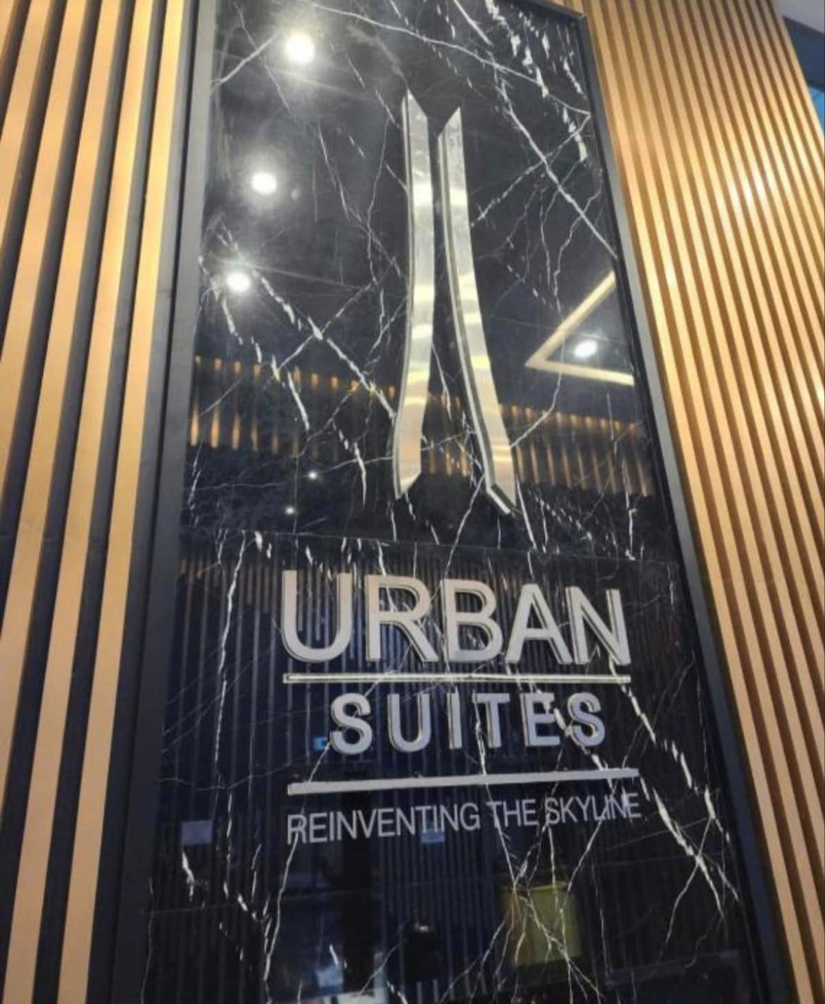 The entrance of Urban Suites is highlighted by a sleek marble wall, featuring the logo in modern typography. Vertical lines are elegantly integrated into the design, complementing the wood paneling that adorns the surroundings. Soft lighting enhances the contemporary atmosphere.