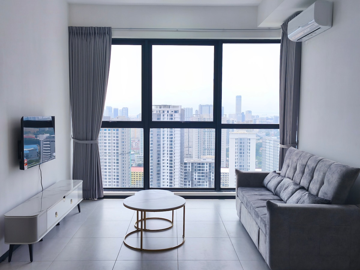 A living area features a comfortable grey sofa and a round coffee table, with a large window displaying expansive views of the city skyline. Air conditioning is installed, and a wall-mounted television is positioned opposite the seating area.