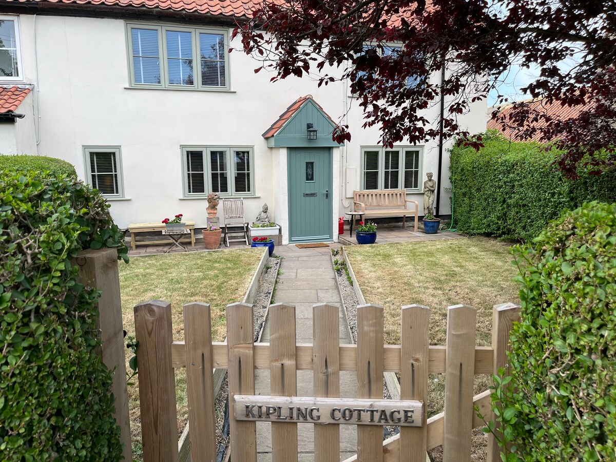 A welcoming entrance is presented with a wooden fence and sign identifying Kipling Cottage. A pathway lined with flowers leads to a charming front door framed by windowed walls. Adjacent seating areas and well-maintained greenery provide a serene outdoor space.