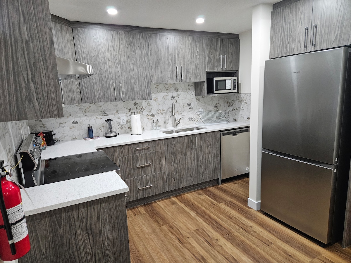 A fully equipped kitchen features modern cabinetry with a wood finish, stainless steel appliances, and an elegant marble backsplash. Essential appliances include a refrigerator, stove, microwave, and dishwasher. The spacious countertop provides ample workspace, complemented by warm wood flooring throughout the area.