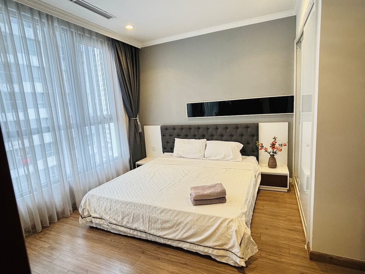 A spacious sleeping area features a comfortable bed with a light-colored blanket. Decorative accents, such as a vase with flowers, are placed on a bedside table. Large windows with sheer curtains allow natural light to fill the room, creating a bright and welcoming ambiance.