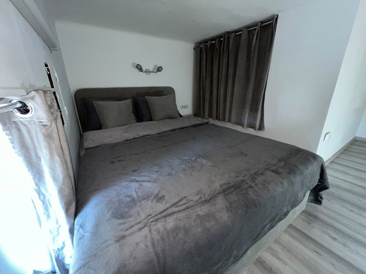 A spacious bed is covered in soft, gray bedding, flanked by two pillows. The room is softly lit, and dark curtains frame a window, allowing natural light to enter.