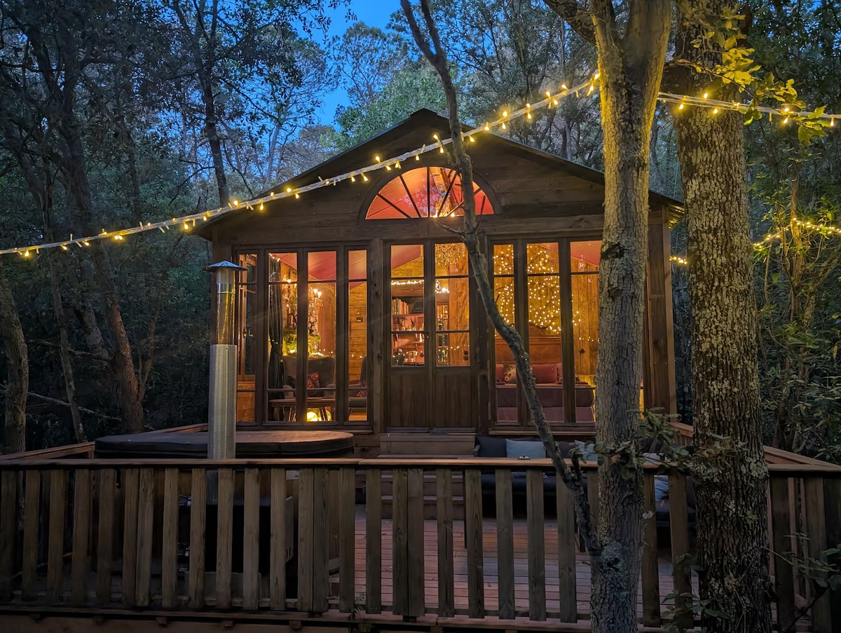 A charming cabin is nestled among the trees, illuminated by soft string lights. Large windows allow a view of the cozy interior, where warmth is provided by a stylish heater. A spacious deck surrounds the cabin, enhancing the immersive forest experience.