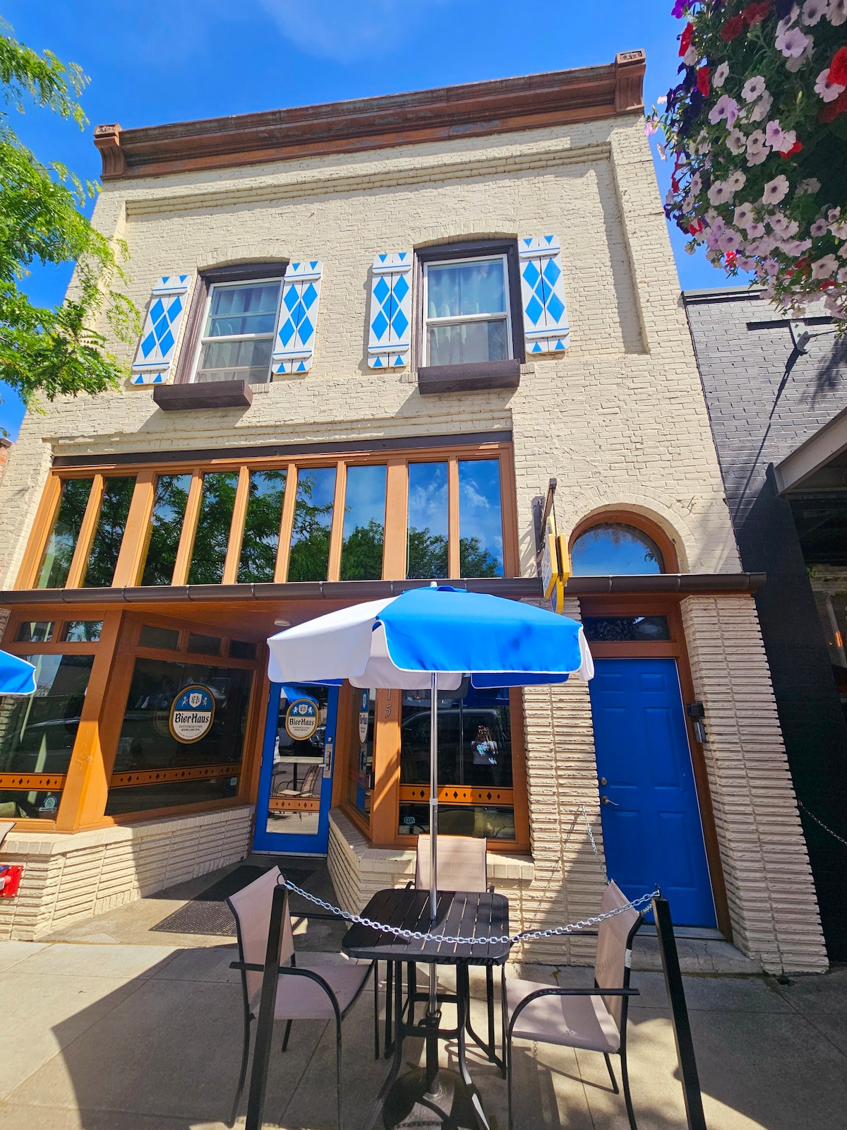 The exterior of a charming building features a bright blue entrance door and large windows framed in wood. Outdoor seating is positioned under a blue and white umbrella on the patio, inviting guests to enjoy the vibrant streetscape of Coeur d'Alene.