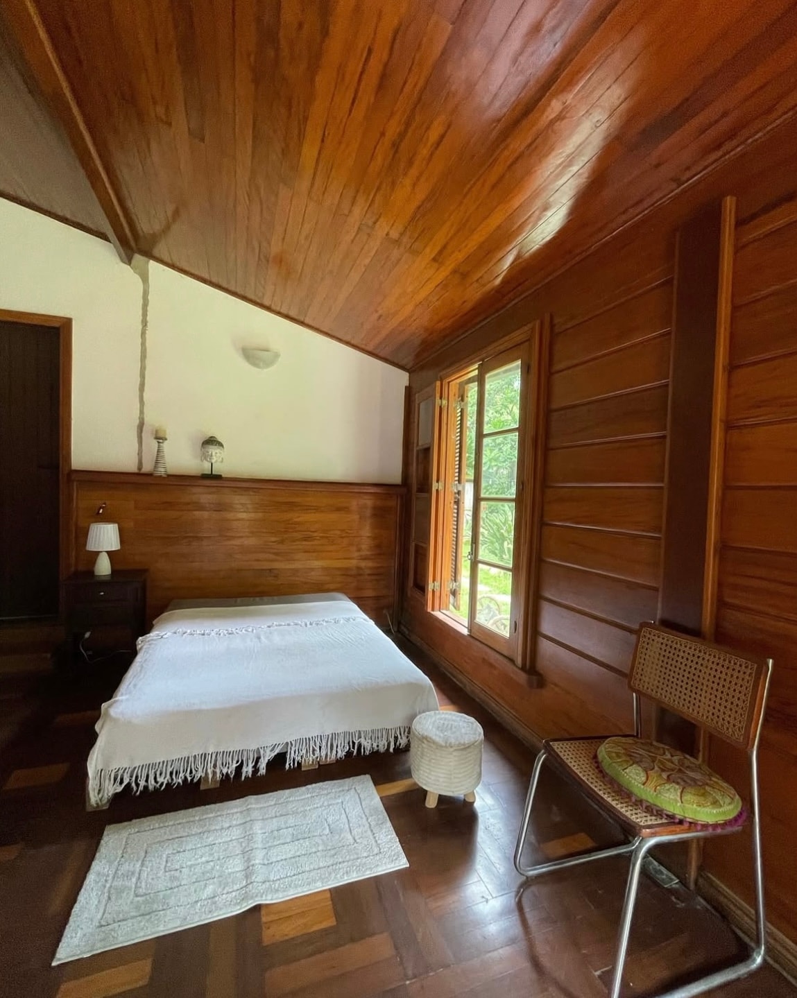 The room features a spacious bed draped with a light blanket, complemented by a subtle rug on the wooden floor. A simple chair is positioned nearby, and natural light filters through wooden-framed windows, creating a serene ambiance with views of the surrounding greenery.