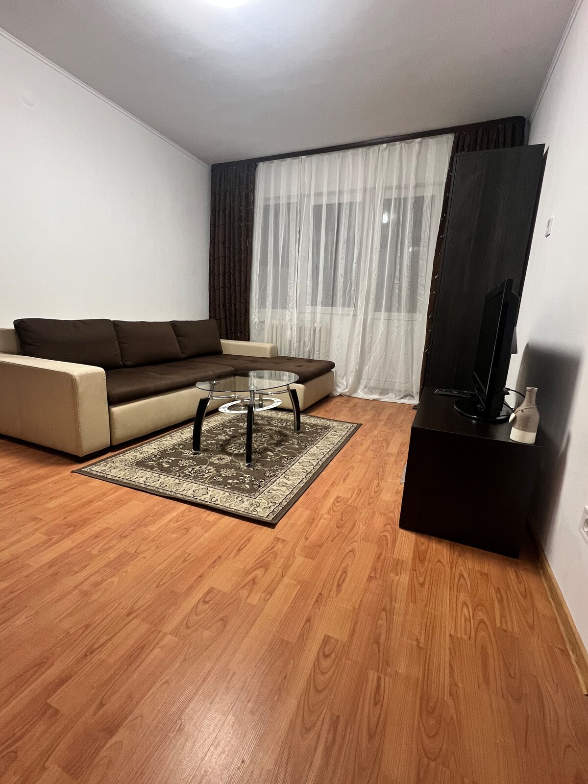 A modern living area features a light-colored sofa paired with a glass coffee table atop a decorative rug. Natural light is allowed in through sheer curtains, illuminating the space. A dark TV unit stands against the wall, complementing the warm tones of the wooden floor.
