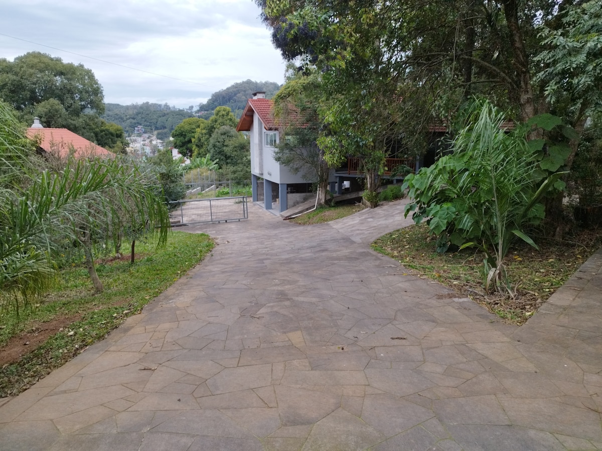 A wide, paved driveway leads to the house, embraced by lush greenery on either side. The surrounding landscape includes various trees and shrubs, contributing to a serene, natural environment. The house is visible in the background, partially hidden among the foliage.