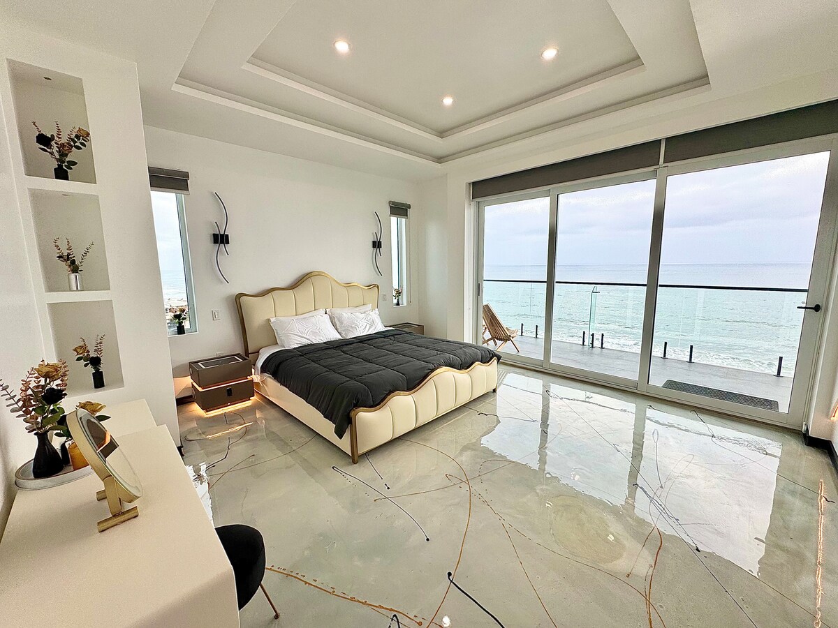 An elegant bedroom showcases a spacious bed with a soft gray comforter. Large glass doors provide an unobstructed ocean view, allowing natural light to fill the room. A dedicated vanity area is positioned beside the bed, while decorative vases add a touch of detail to the space.