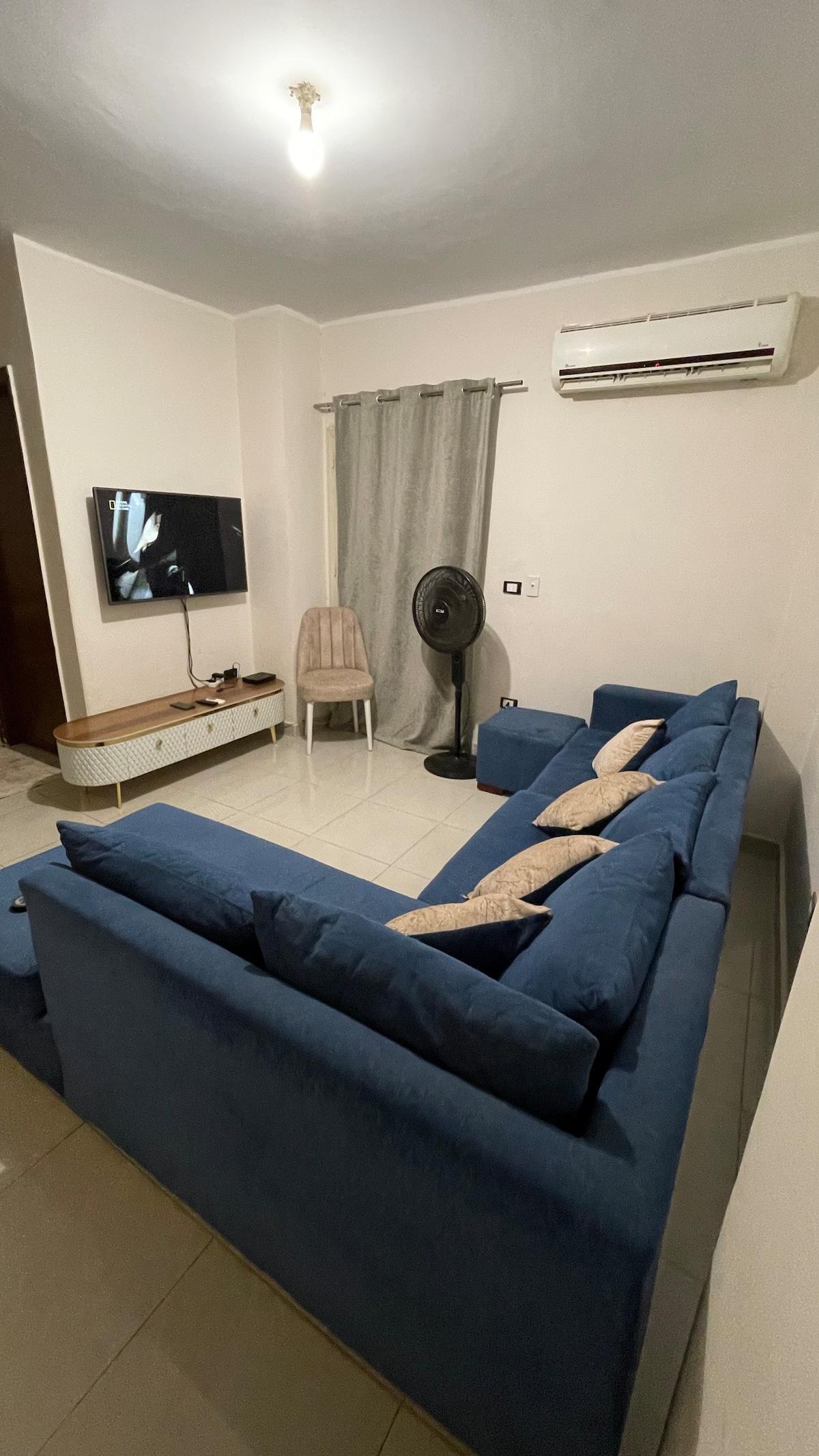 A cozy living room is presented with a blue sectional sofa and a wooden TV stand. A flat-screen TV is mounted on the wall opposite the seating area. Natural light filters in through a window, with a curtain covering it partially. A decorative chair and a fan complete the space.