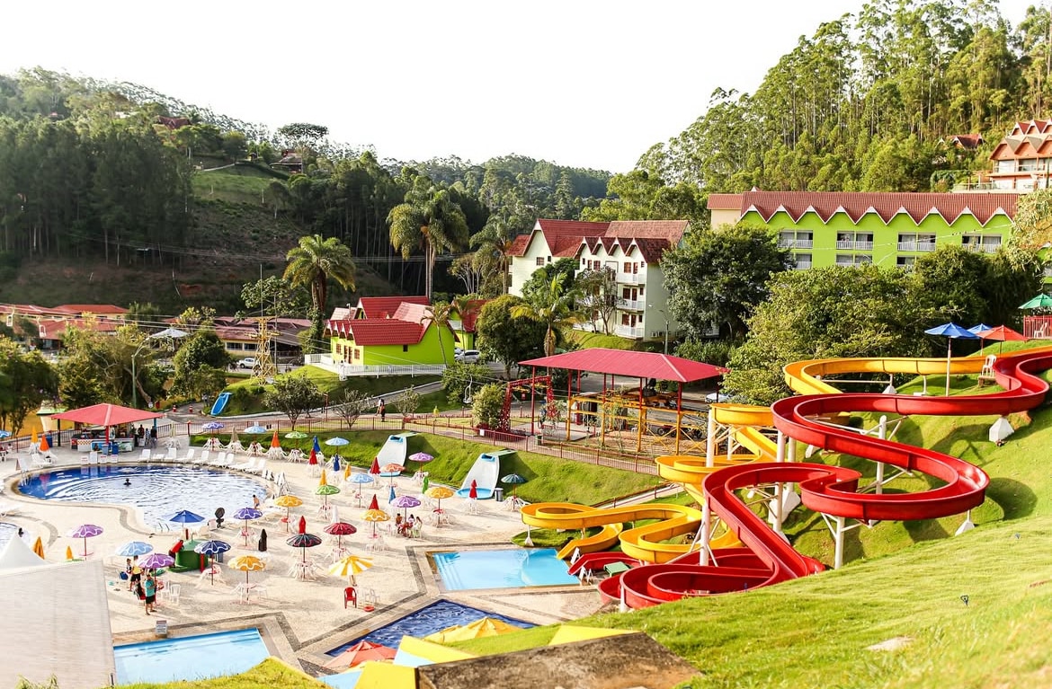 A vibrant water park area is highlighted, featuring colorful water slides in red and yellow. Several swimming pools are visible, surrounded by lounge chairs and umbrellas. The resort buildings with green and red accents are set against a backdrop of lush hills and trees.