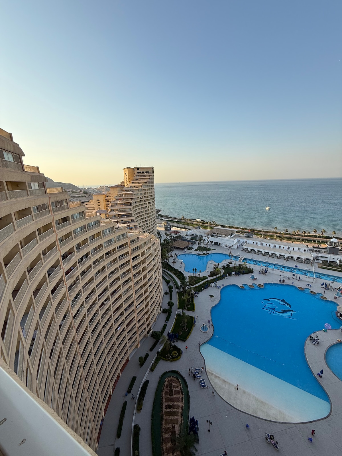 The image captures a panoramic view of a resort complex by the sea. Curved architectural designs of high-rise buildings are visible, overlooking a large, serene swimming pool. Surrounding greenery and walkways lead toward the beach, with the horizon blending into a clear evening sky.