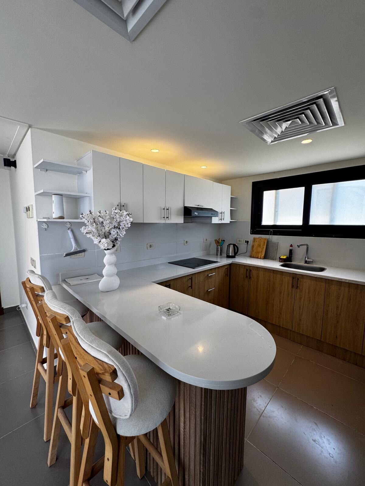 The kitchen area features a modern design with a curved countertop and wooden accents. High-backed stools are positioned at the counter. Light cabinets are complemented by a range hood and sleek appliances, with ample natural light coming through large windows.