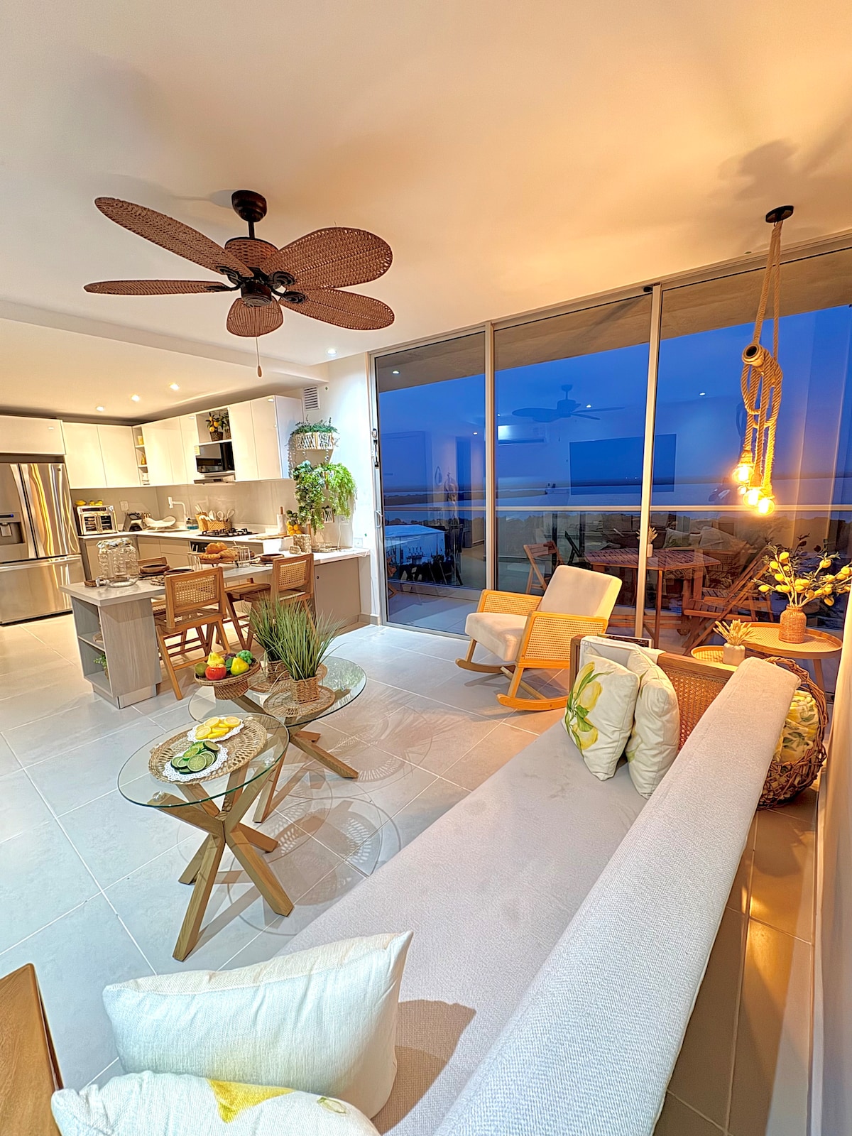 A bright living space is depicted, showcasing a comfortable sofa paired with two chairs. Natural light floods in through large windows. A ceiling fan adds a cooling element, while a dining area is visible in the background, featuring wooden furniture and a well-equipped kitchen.