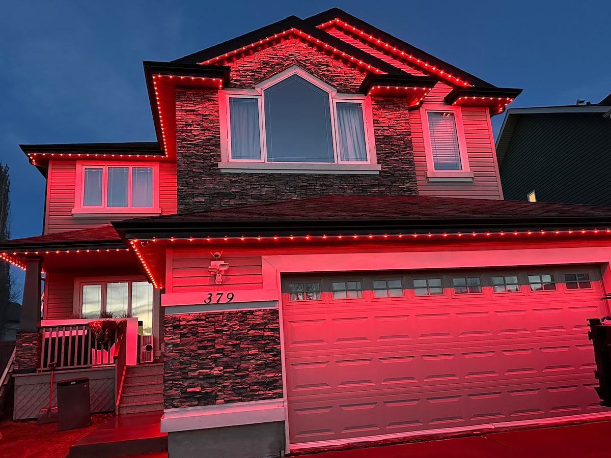 A two-story house is illuminated by red LED lights outlining the roof and windows. The stone-clad facade contrasts with the vibrant lighting, while a garage door is visible, partially reflecting the glow. Large windows allow for natural light, framed by curtains.