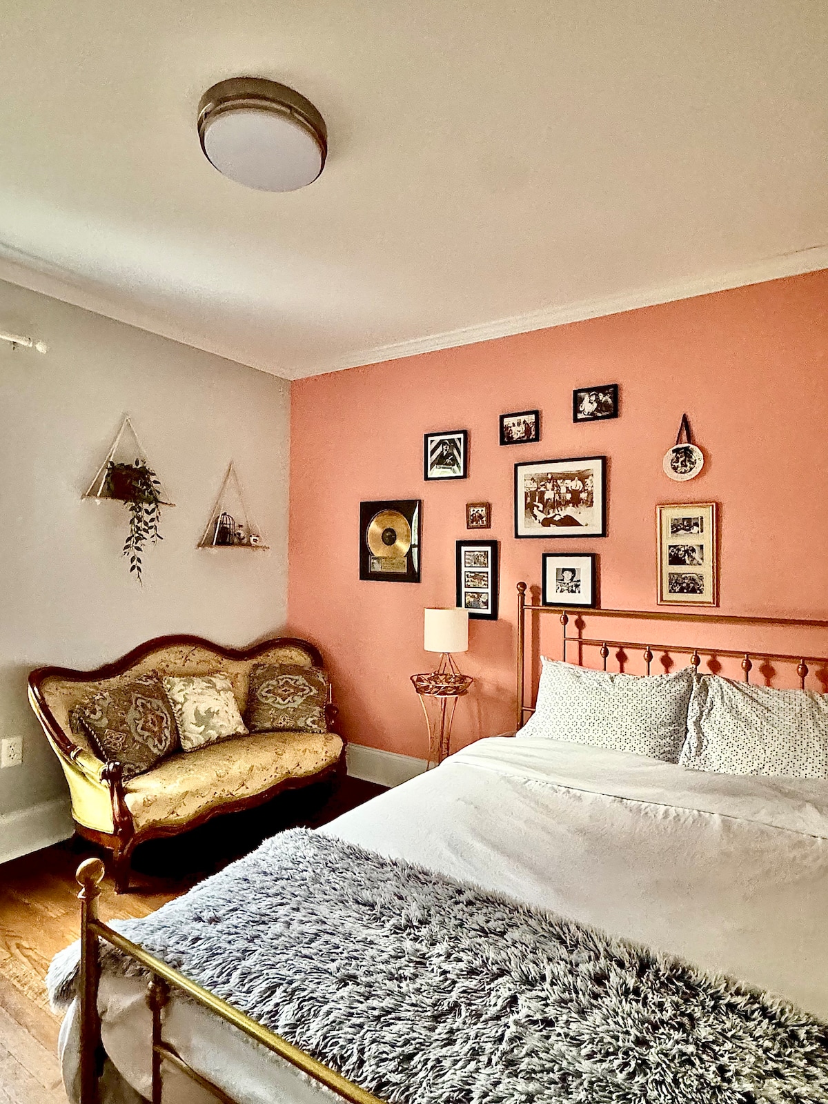 A cozy bedroom is presented with a metal bed frame and plush bedding. A vintage-style sofa is positioned beside the bed, accompanied by a lamp on a small table. The wall features an array of framed photos and decorative elements, enhancing the inviting atmosphere.