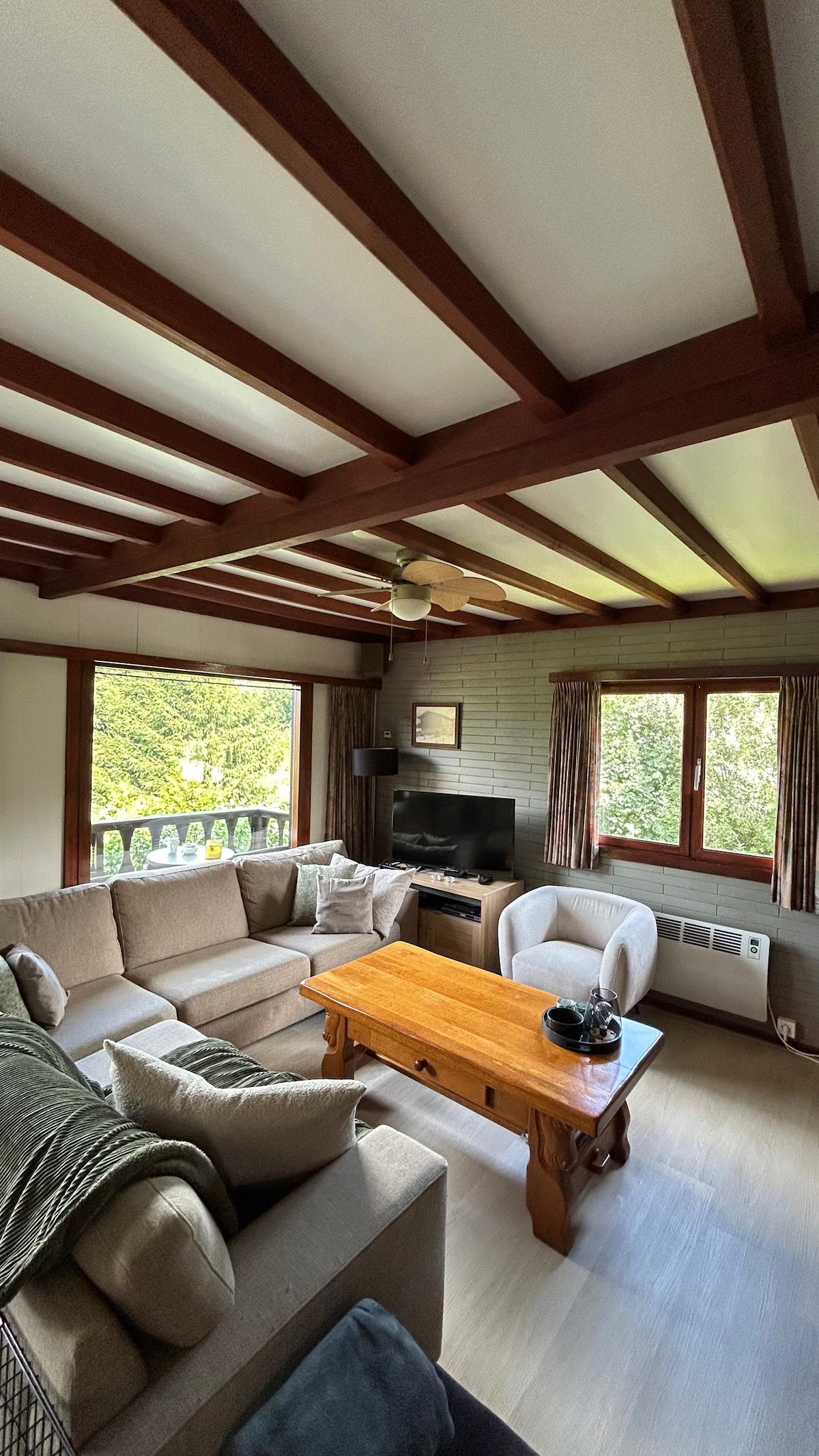 A comfortable living area features a spacious sectional sofa facing a wooden coffee table. Natural light filters through large windows, revealing views of the outdoors. The room is complemented by a television and neutral decor, inviting relaxation and conversation.