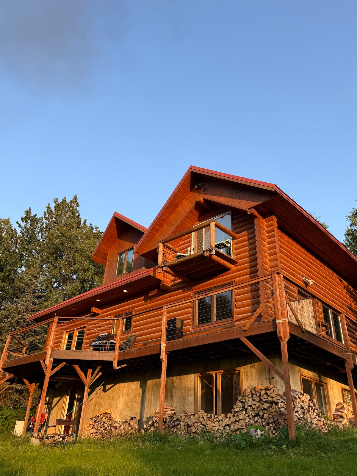 The rustic log home features a striking red roof and multiple levels, with a spacious balcony extending from the upper level. Stacked firewood is neatly arranged beneath the porch, surrounded by lush green grass and trees, creating a natural setting.