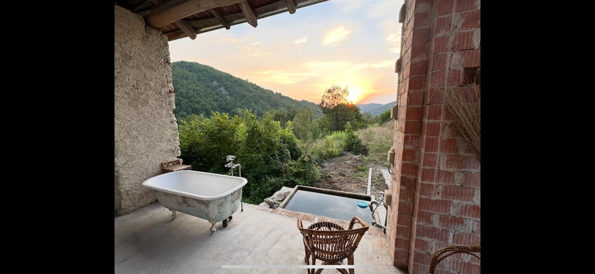 An outdoor bathing area is presented, featuring a vintage bathtub and a plunge pool with natural surroundings. A wicker chair is positioned nearby, with a view of rolling hills and a sunset illuminating the sky, creating a serene atmosphere.