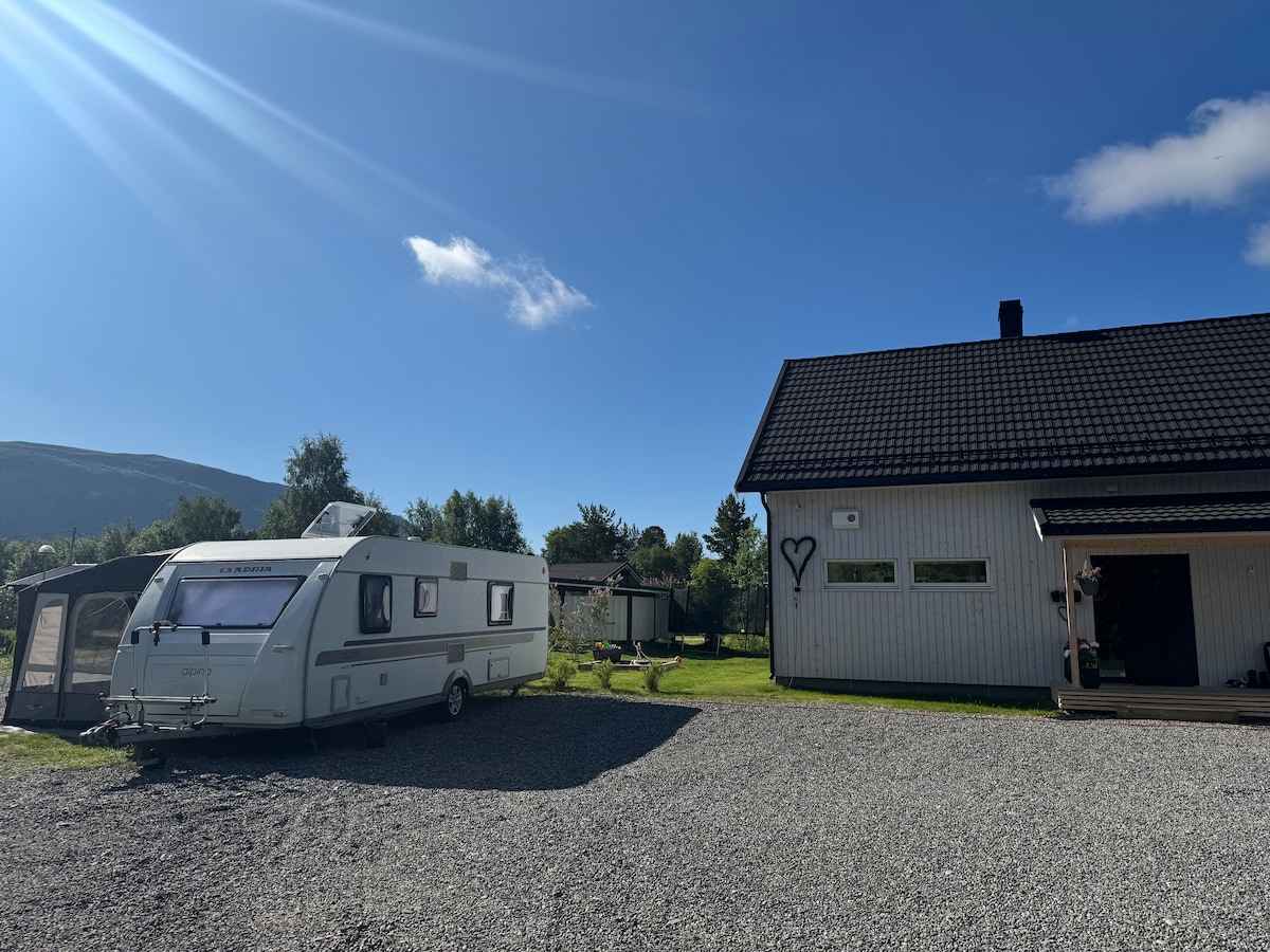 A caravan is parked on a gravel area in front of a modern, light-colored house. The sky above is clear with a few clouds and sunlight casting gentle shadows. Lush greenery surrounds the setting, creating a serene environment.