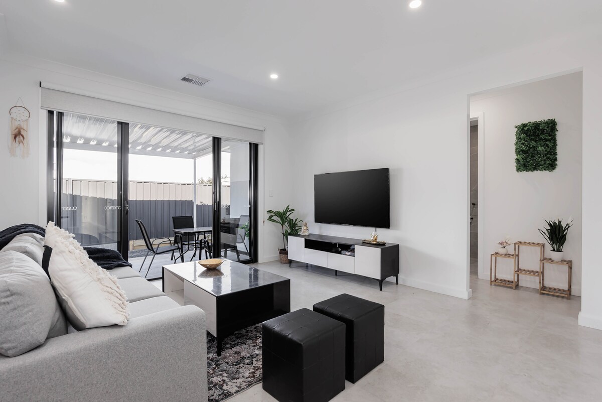 A modern living room features a comfortable gray sofa accompanied by two black ottomans. A sleek coffee table sits in the center, with a television mounted on the white cabinetry. Large glass sliding doors open to a patio area, allowing natural light to fill the space.