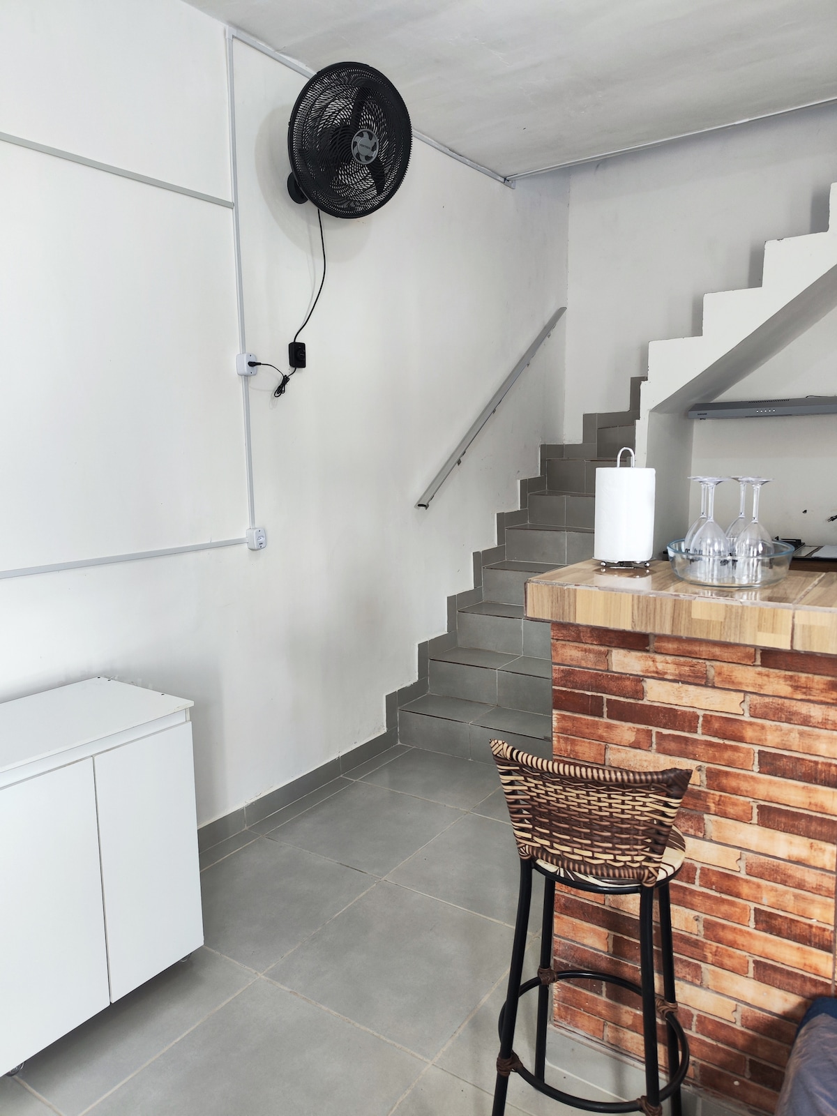 An indoor space features a staircase leading to the upper level, with a neutral palette and modern design. A bar area with a wooden countertop is accompanied by a woven stool. A wall-mounted fan provides ventilation, enhancing the comfort of the environment.