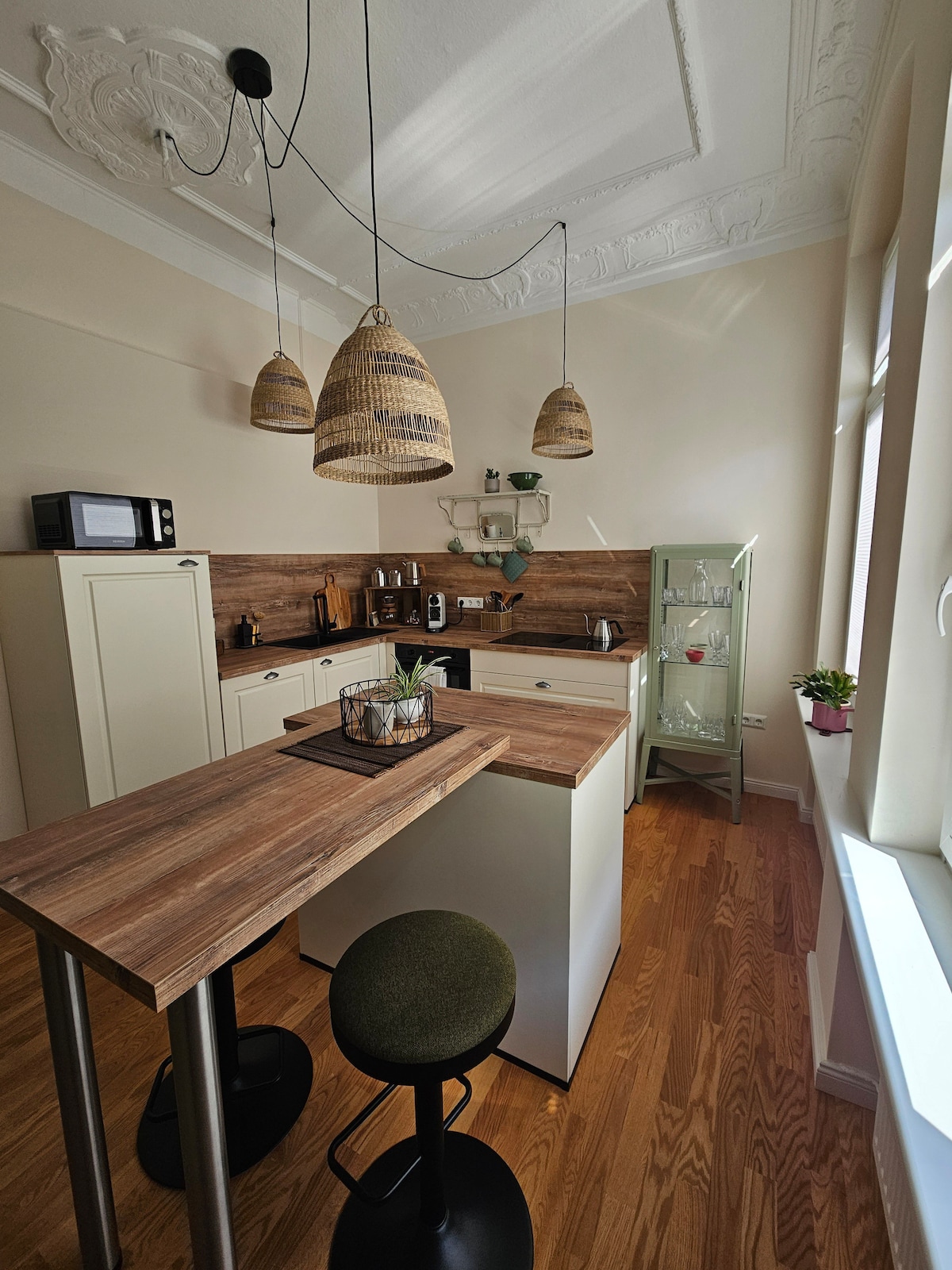 A kitchen area features light wooden cabinetry and a bar-height table with green stools. Three woven pendant lights hang from the ceiling. A microwave and various kitchen appliances are visible, alongside a glass cabinet displaying dishware. Natural light streams through two large windows.