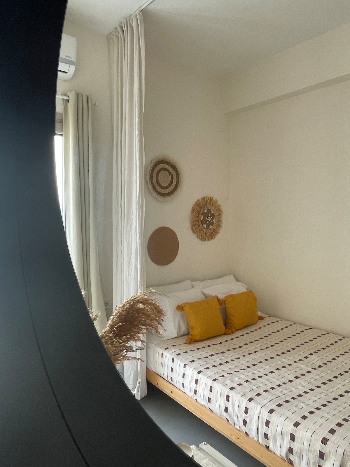 A spacious bed is positioned against a neutral-colored wall, adorned with decorative elements like woven wall art. Soft pillows are arranged neatly at the head of the bed, while light filtering through curtains adds a calming ambiance to the room.