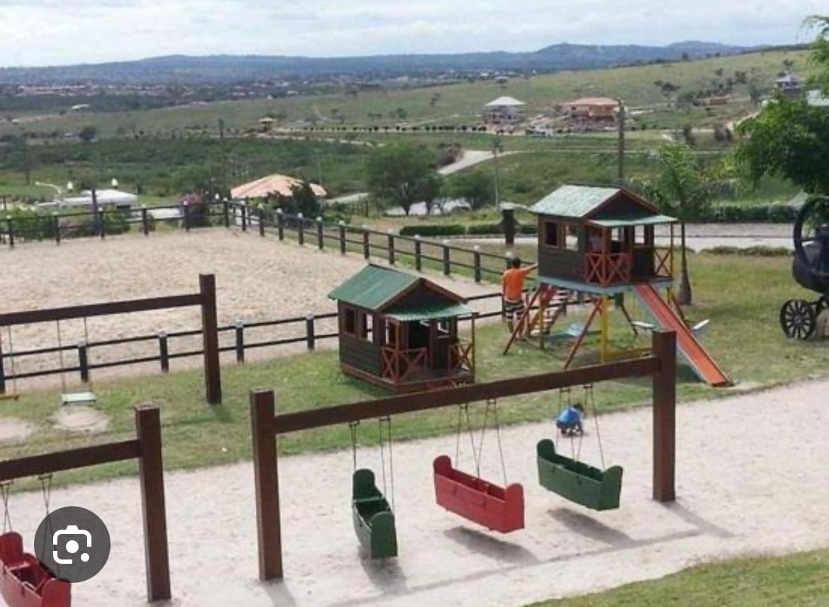 A children's play area is depicted, featuring two wooden playhouses and a slide. Nearby, a row of swings in vibrant colors is available for use. The surrounding landscape includes a gently rolling terrain and distant structures, contributing to an open, outdoor setting.