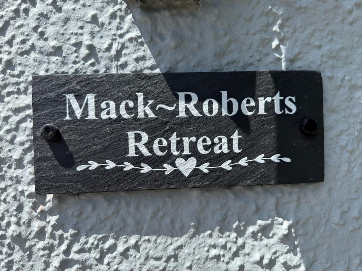 A decorative slate sign displays the name 'Mack-Roberts Retreat,' accentuated by a simple heart and floral motif. The sign is fixed to a textured white wall, providing clear visibility of its bold lettering.