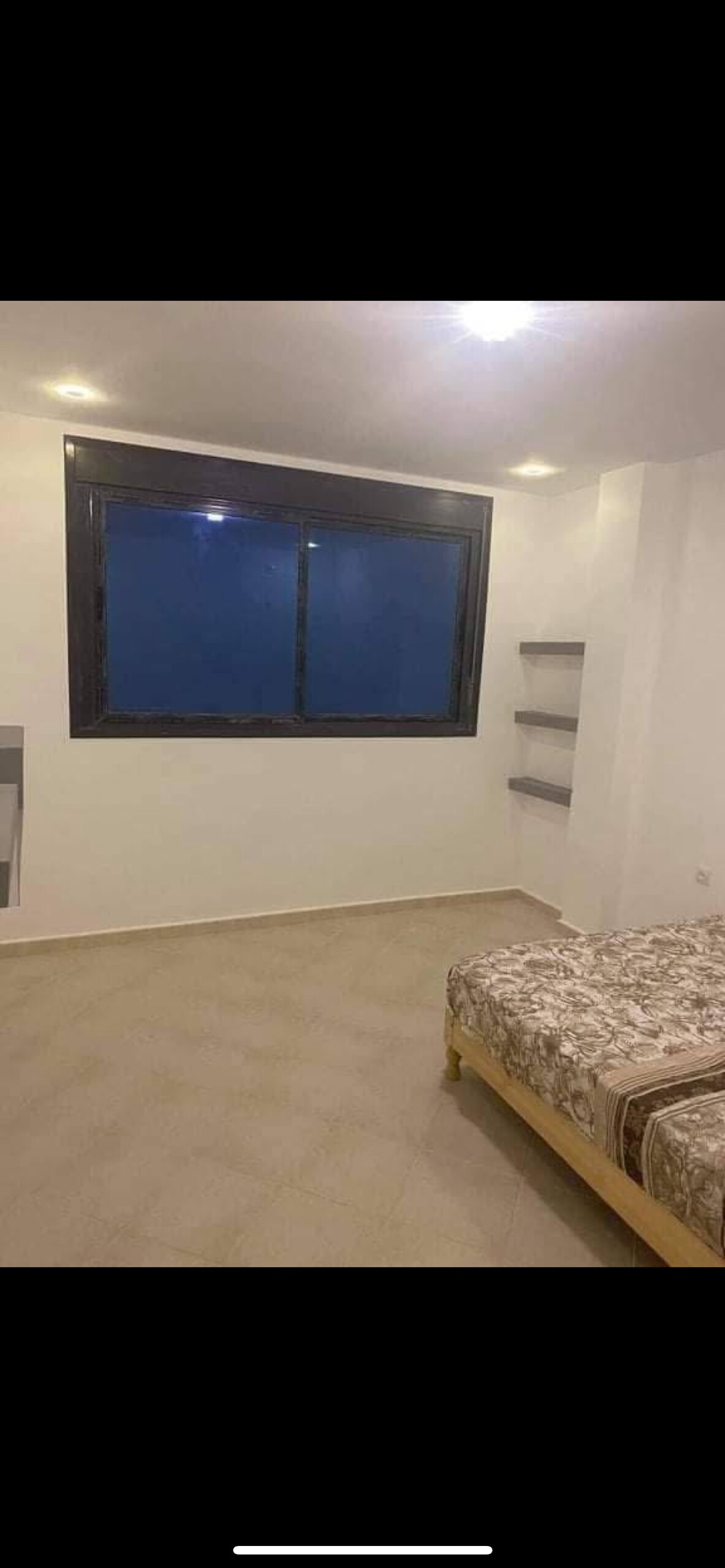 A spacious bedroom features a large window allowing natural light to fill the room. Two beds with patterned bedding are positioned against one wall. Shelves are mounted on the opposite wall, offering additional storage or display options.