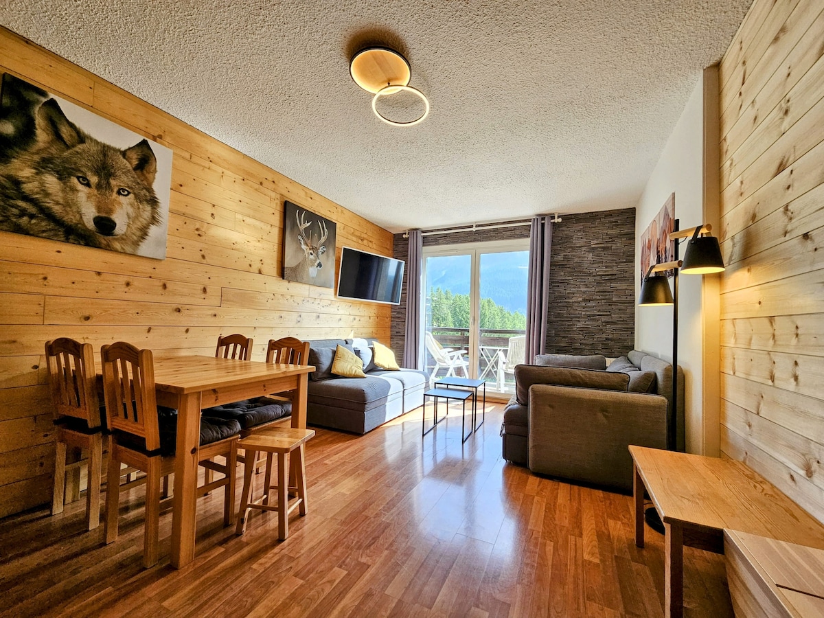 A spacious living area features wooden paneling and a large window overlooking the mountains. A dining table with four chairs sits beside a cozy sofa and an armchair, accompanied by a coffee table. Artwork depicting wildlife adorns the walls, enhancing the ambiance.