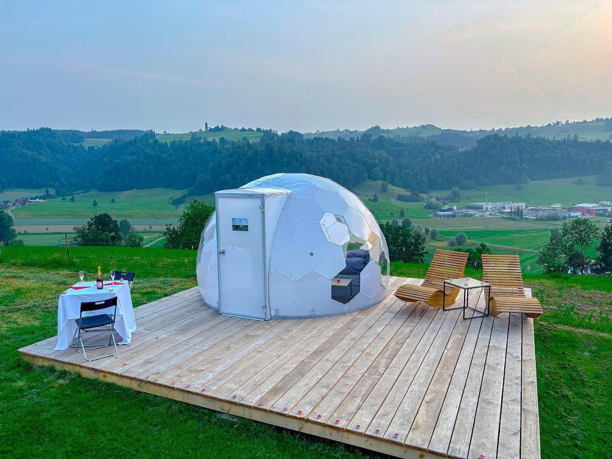 A unique geodesic dome tent is positioned on a wooden deck, surrounded by grassy fields and rolling hills. Two lounge chairs are placed nearby, along with a small table set for two, creating a serene outdoor setting for relaxation and dining.