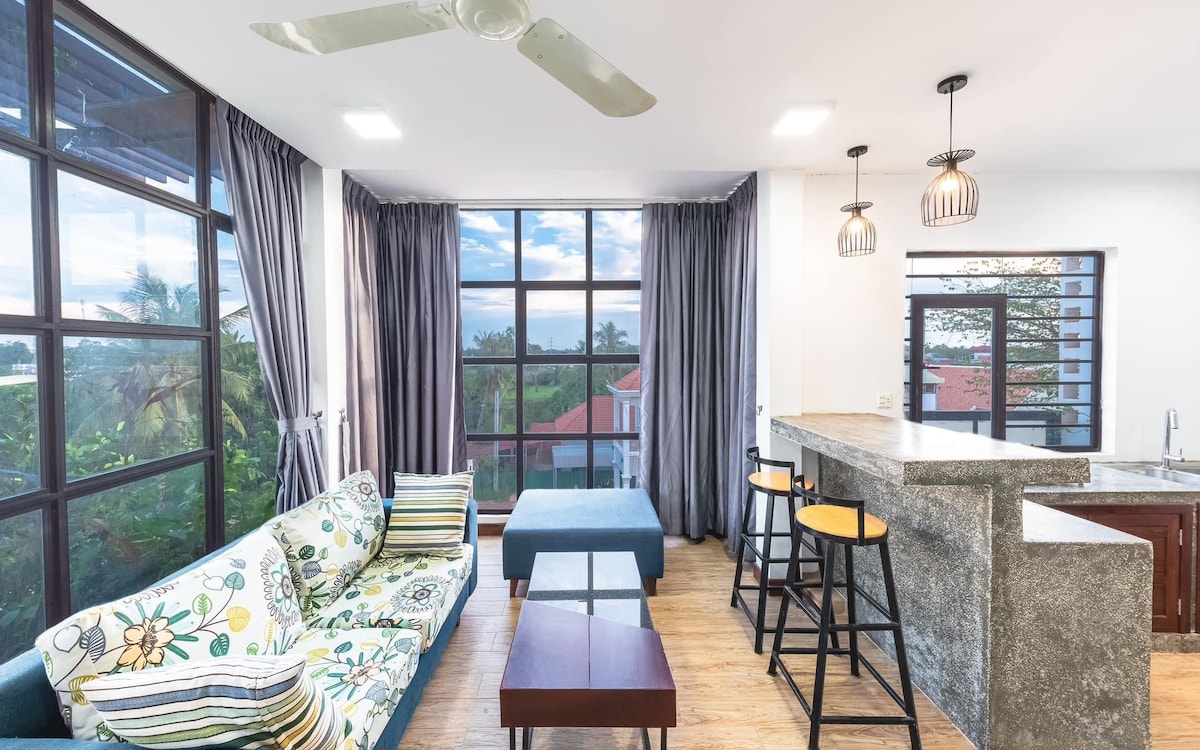 A modern living area features large windows allowing natural light to fill the space. A floral-patterned sofa and a blue ottoman provide seating options, while a breakfast bar with four stools is positioned next to a kitchen counter. Light-colored curtains frame the windows.