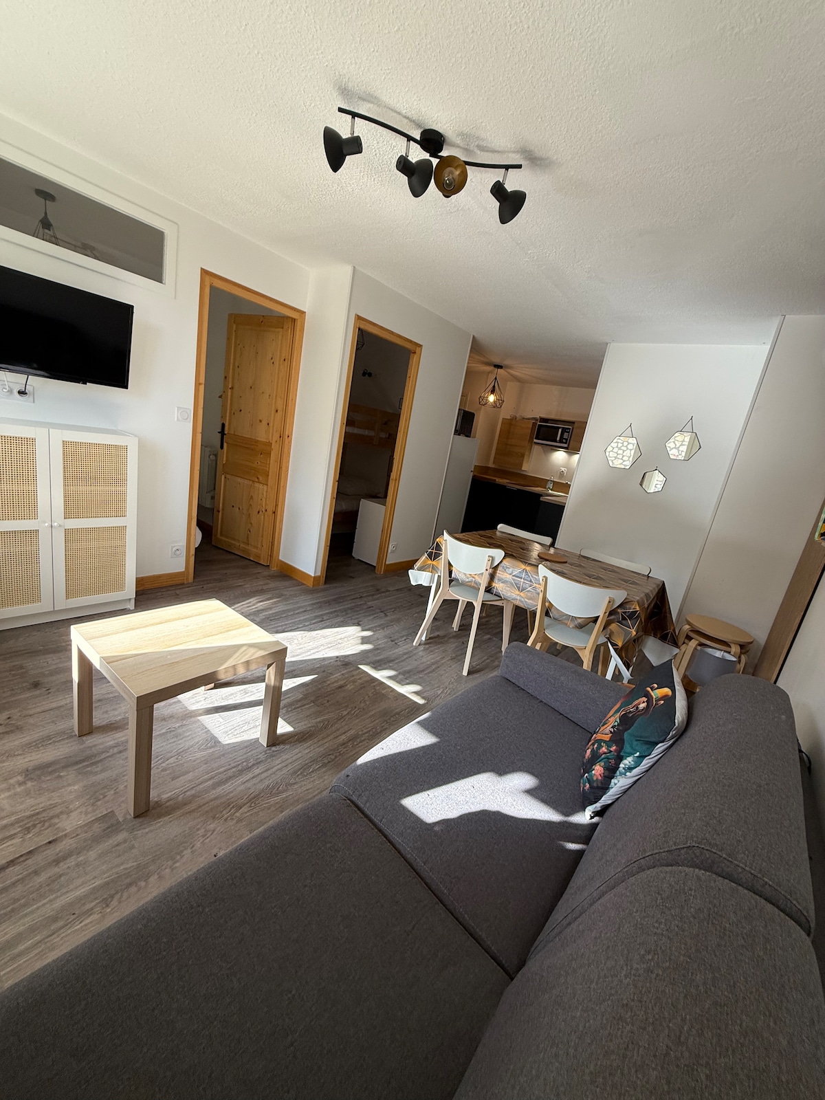 A spacious living area is showcased, featuring a gray sofa and a light wooden coffee table. A dining set with four white chairs surrounds the table, and a wall-mounted TV is visible. The kitchen area is partially seen, indicating a practical layout. Natural light enters from an unseen window.