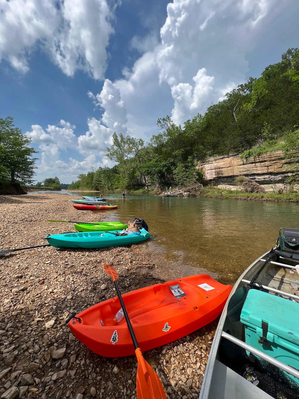 Colorful kayaks are lined up along a rocky creek bank, featuring bright orange, green, and blue hues. The clear waters reflect the surrounding greenery and distant rock formations, while fluffy clouds fill the blue sky, creating a peaceful outdoor setting for water activities.