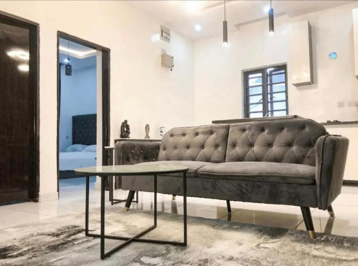 A modern living area is showcased, featuring a tufted gray sofa opposite a glass coffee table. The room is illuminated by pendant lights, and a cozy rug lies beneath. A doorway leads to a separate sleeping space, visible in the background.