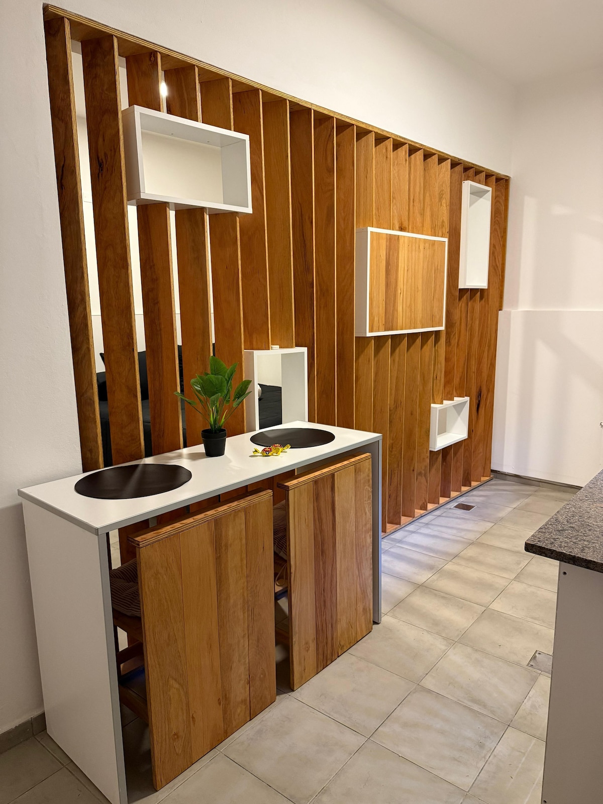 A sleek dining area features a minimalist design with a light countertop and two round sinks. Wooden elements provide a warm contrast, complemented by decorative shelves on the wall. A small potted plant adds a touch of greenery to the space, enhancing the modern aesthetic.