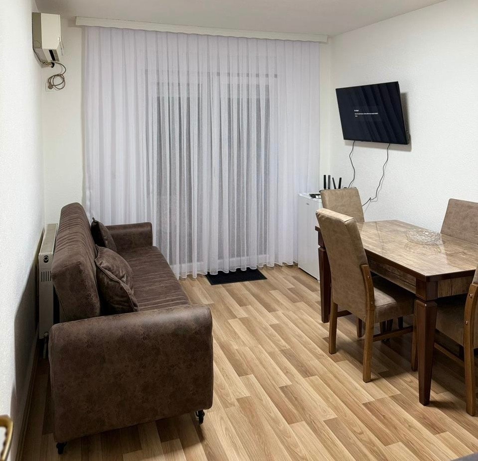 A living area features a brown sofa in front of a wall-mounted television. A dining table is positioned beside four cushioned chairs. Natural light enters through sheer white curtains, illuminating the light wood floor.