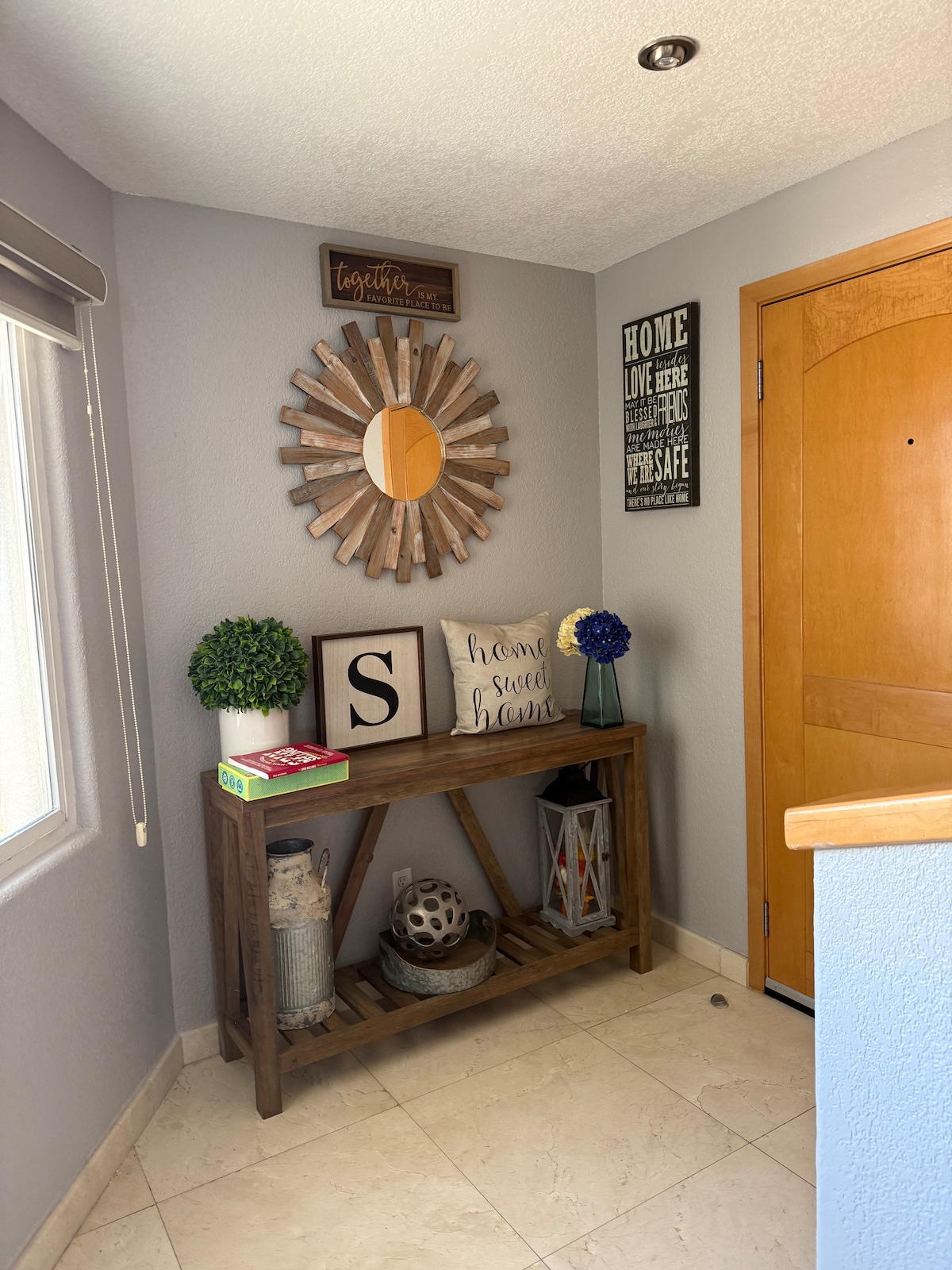 An entryway features a wooden console table positioned against a wall. A round mirror hangs above the table, reflecting light. Decorative items include a blue flower arrangement, framed letters, and a lantern. A wooden door with a round handle stands to the right.