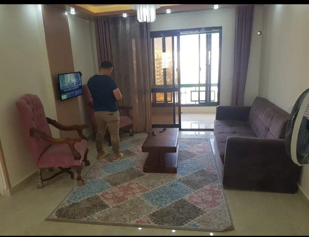 A living area is shown, featuring a brown sofa and two armchairs, one in a soft pink hue. A coffee table is positioned centrally on a patterned rug. Light flows in through large glass doors, leading to a balcony with city views.