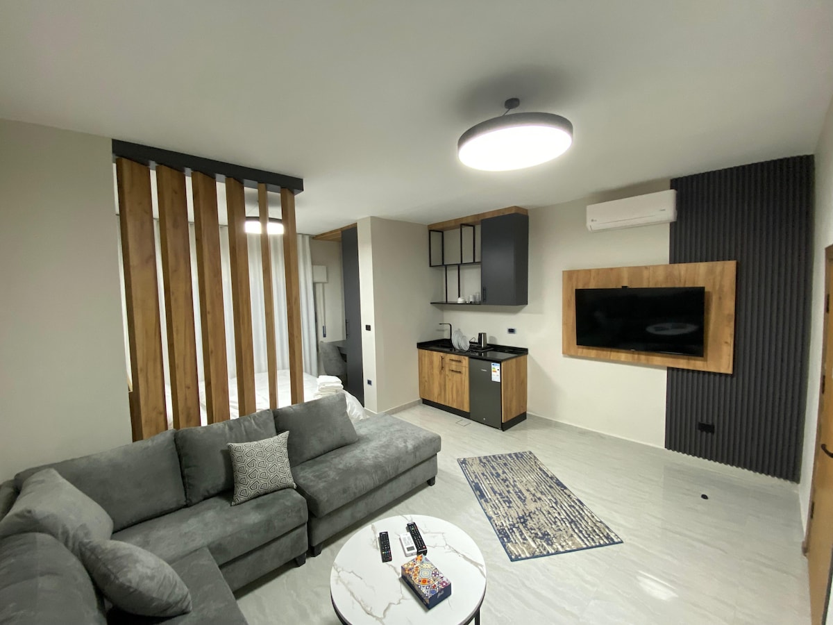 A contemporary living area features a comfortable grey sectional sofa, paired with a round coffee table. A flat-screen TV is mounted on the wall, while a kitchenette with dark cabinetry is visible in the background. Light from a ceiling fixture enhances the overall ambiance.