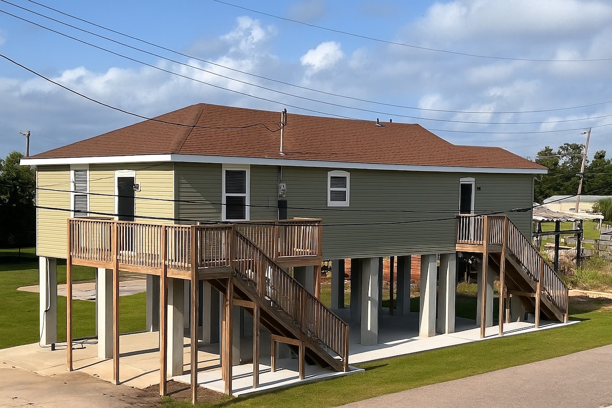 The elevated bayou home features a two-story structure with a light green exterior and a red-brown roof. Two sets of wooden stairs provide access to the upper level, while a spacious area is visible underneath the house, surrounded by a grassy yard.